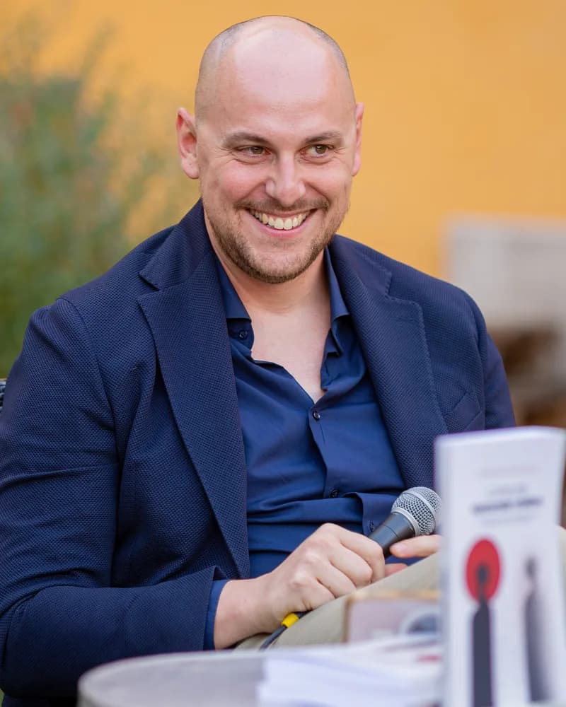 Gabriele Pecchioli sorride seduto, con il microfono in mano e una copia del suo libro sul tavolino